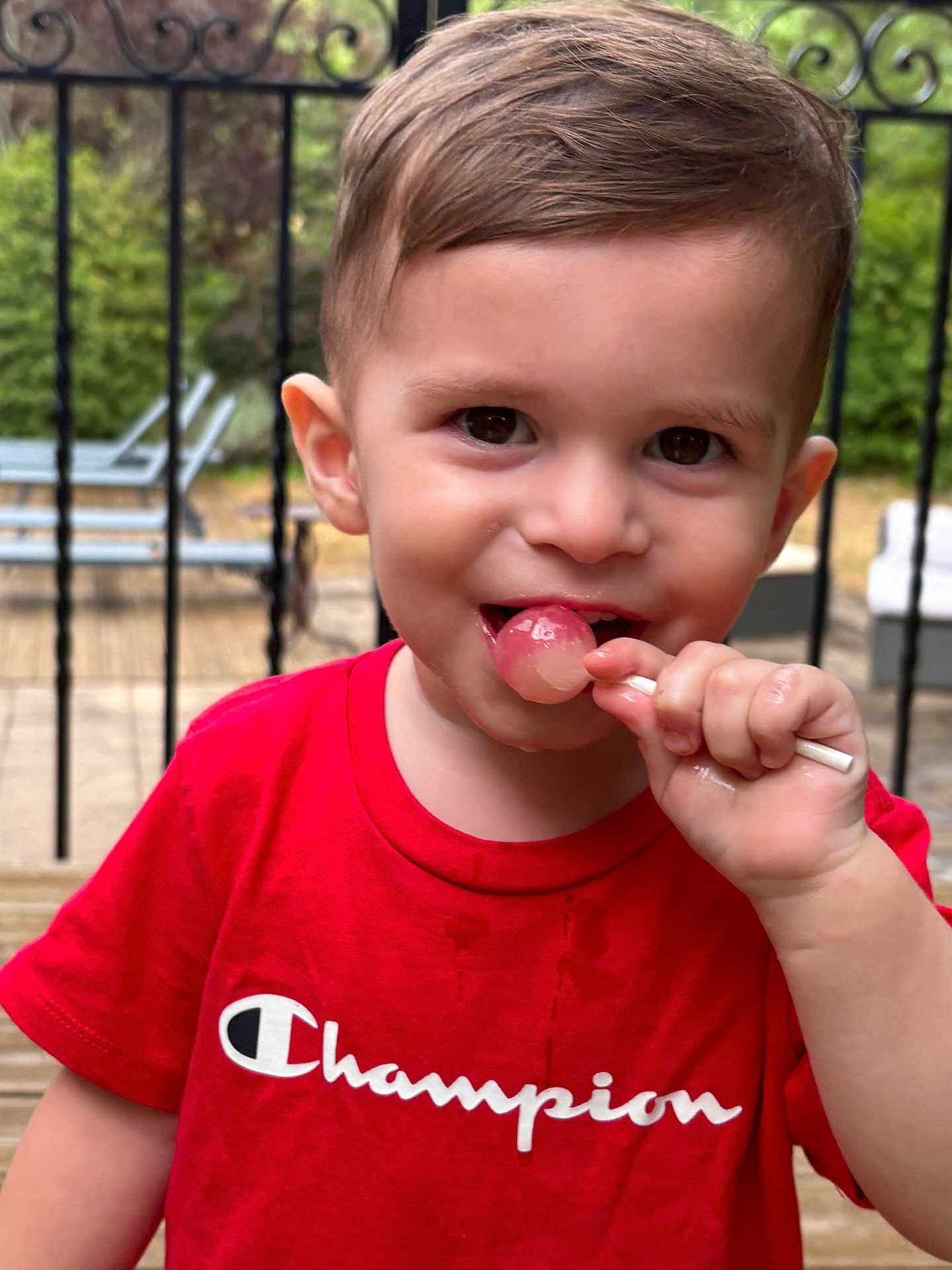 Soan participe au concours pour gagner de l'argent avec cette photo : toddler, child, red_shirt, lollipop, smiling, outdoor, deck, wooden_floor, garden, patio_furniture, boy, short_hair, daylight, happy, cute, person, portrait, summer, playful, casual_clothing