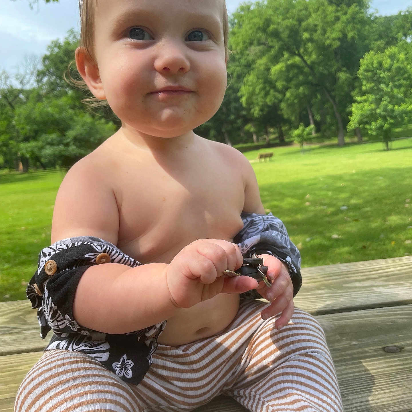Kesston is registered to the contest to win money with this photo: baby, bench, child, cute, daytime, grass, greenery, holding_object, nature, outdoor, park, person, playing, shirtless, smiling, striped_pants, sunlight, toddler, trees, wooden_table