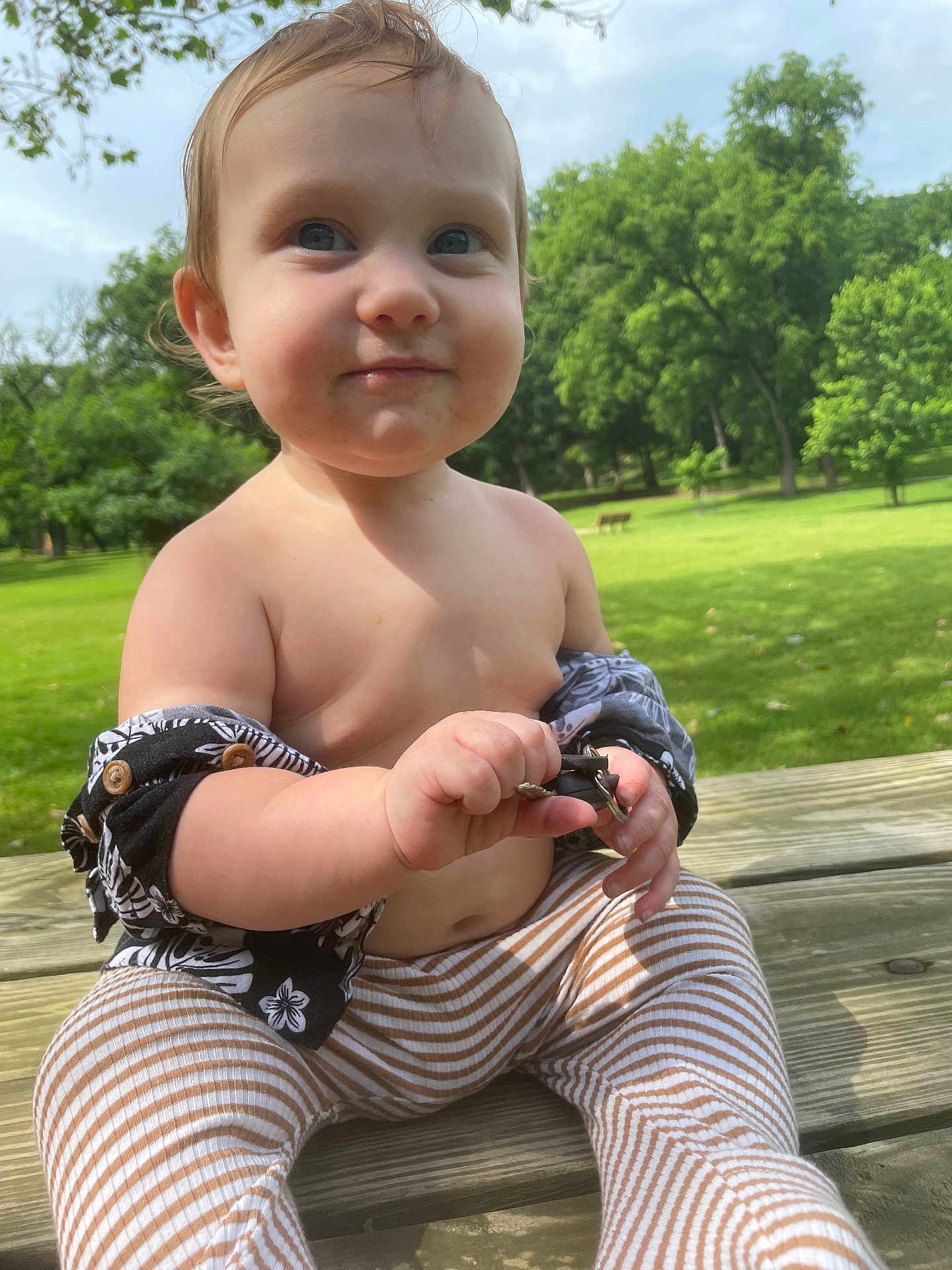 Kesston is registered to the contest to win money with this photo: toddler, child, outdoor, park, grass, trees, bench, shirtless, striped_pants, baby, smiling, wooden_table, nature, sunlight, greenery, person, cute, playing, holding_object, daytime