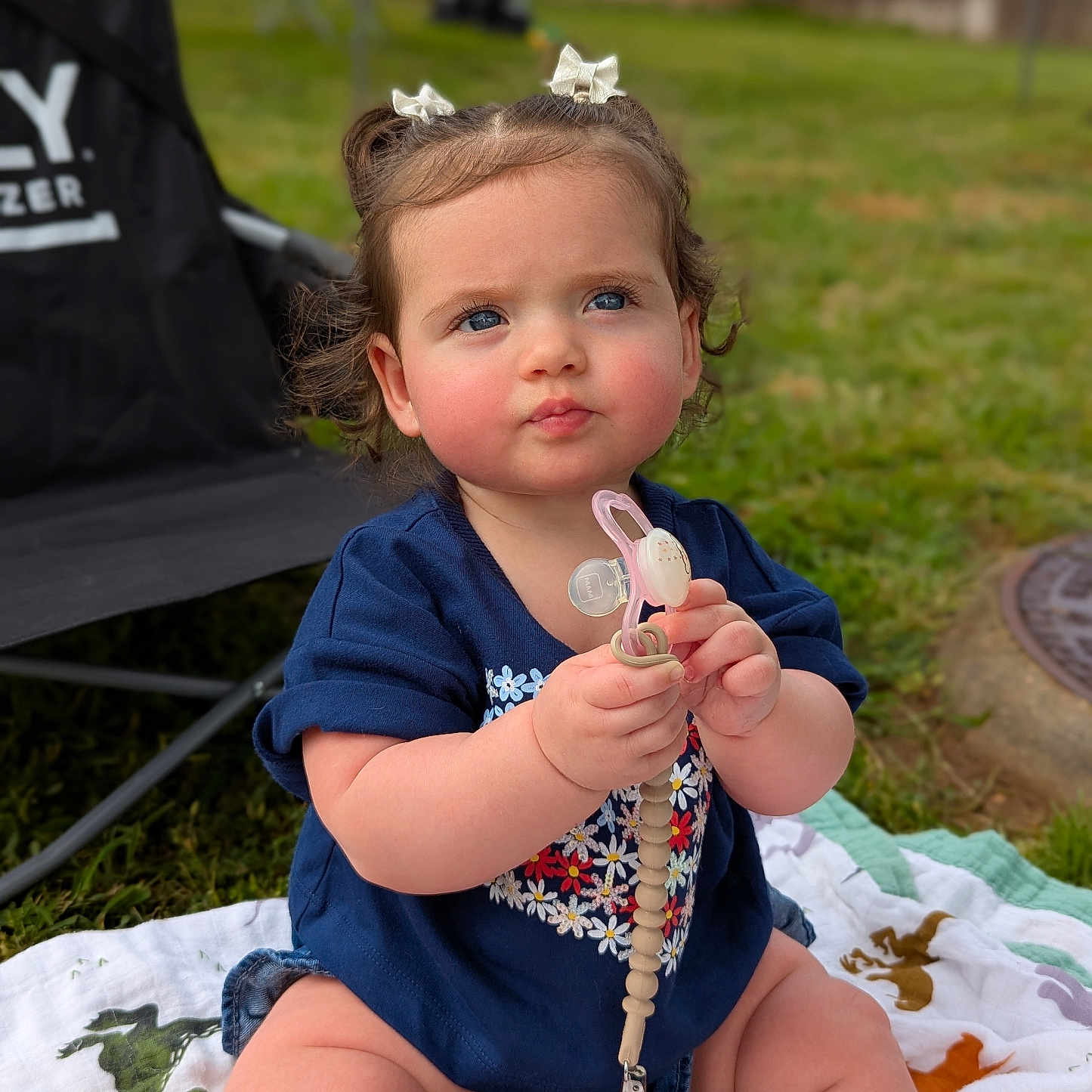 Amelia is registered to the contest to win money with this photo: baby, blanket, blue_eyes, bows, chair, child, clothing, curious, cute, grass, infant, nature, outdoor, pacifier, person, portrait, shirt, shorts, sitting, toy