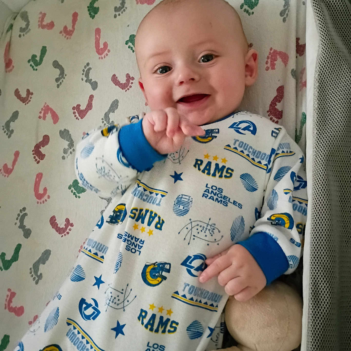 Noah joined the competition — help win amazing prizes! baby, infant, smiling, onesie, plush_toy, blanket, footprint_pattern, bed, laying_down, happy, child, cute, blue_cuffs, los_angeles_rams, toy, soft_toy, infant_clothing, face, hand, indoor