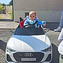 baby, child, toy_car, audi, e_tron_sportback, pacifier, blue_clothing, outdoor, sunlight, sidewalk, house, window, fence, asphalt, person, footwear, shadow, seatbelt, driving, cute