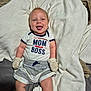 baby, infant, smiling, tongue_out, clothing, mittens, blanket, bed, cute, happy, person, child, apparel, socks, lying_down, indoor, portrait, young_child, text_on_clothing, fabric