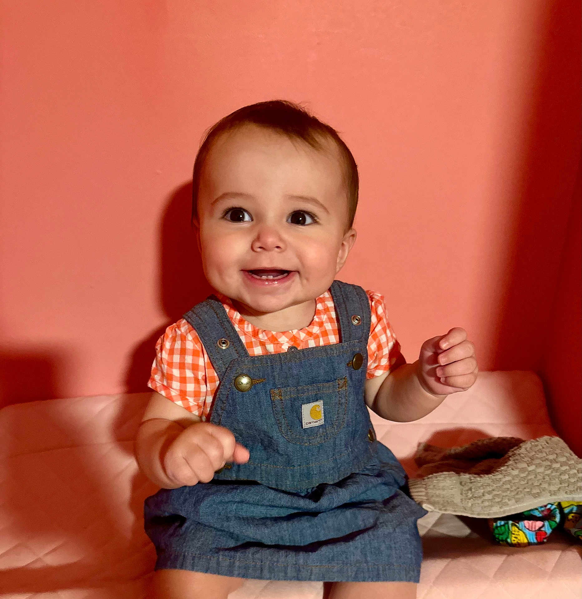 Clover joined the competition — help win amazing prizes! baby, child, smiling, denim_overalls, orange_shirt, sitting, indoor, face, cute, happy, infant, portrait, clothing, human, pink_background, hands, eyes, cheeks, tooth, baby_girl
