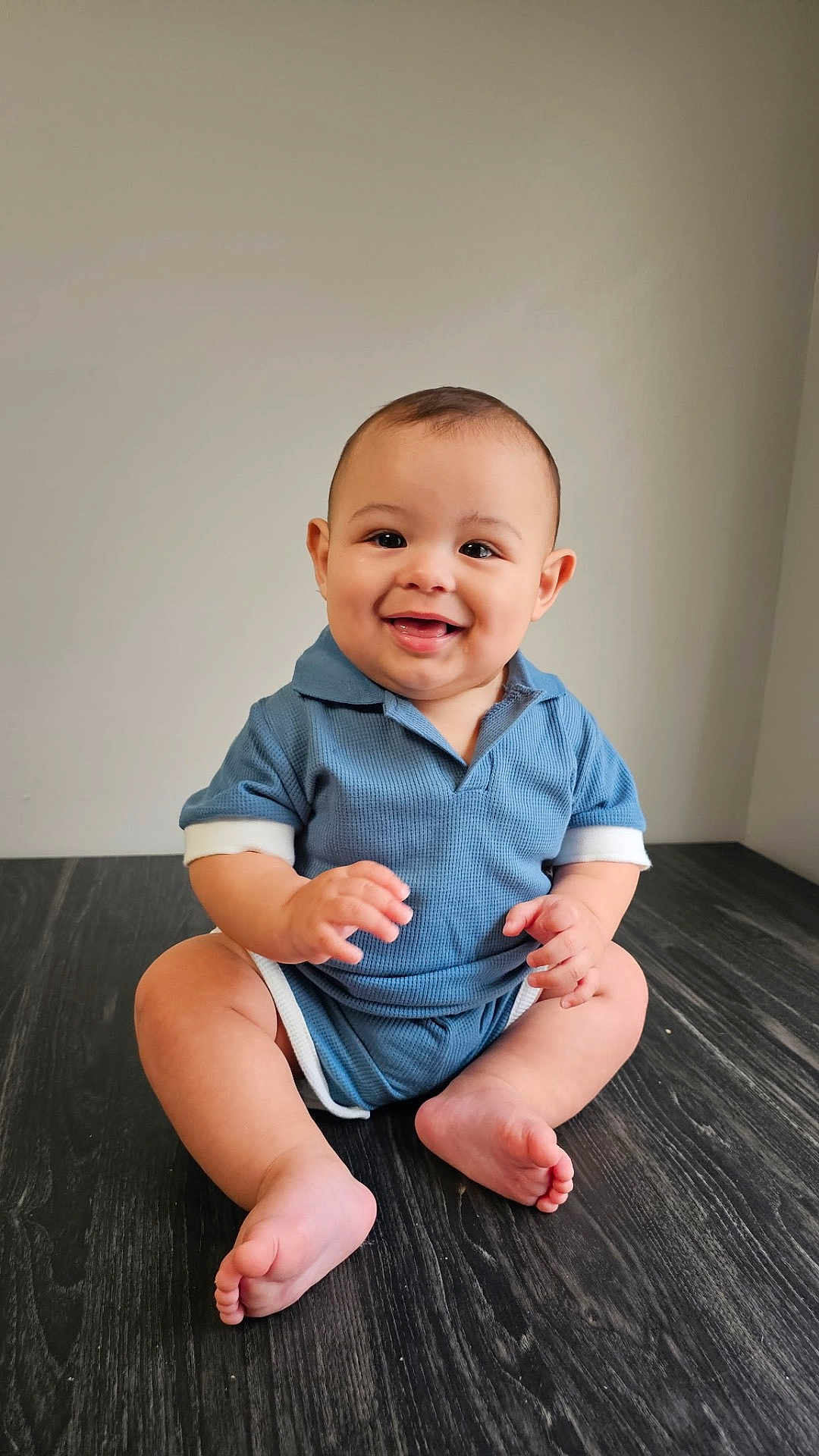 Cooper is registered to the contest to win money with this photo: baby, child, smiling, blue_clothing, sitting, floor, wooden_floor, indoors, cute, happy, hands, feet, portrait, one_person, infant, young_child, casual_clothing, short_hair, skin, room