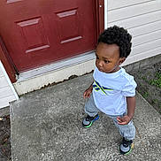 Messiah is registered to the contest to win money with this photo: toddler, child, boy, standing, concrete, door, red_door, white_tshirt, gray_pants, sneakers, outdoor, porch, sidewalk, curly_hair, expression, building, wall, green_plants, casual_clothing, person