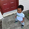 toddler, child, boy, standing, concrete, door, red_door, white_tshirt, gray_pants, sneakers, outdoor, porch, sidewalk, curly_hair, expression, building, wall, green_plants, casual_clothing, person