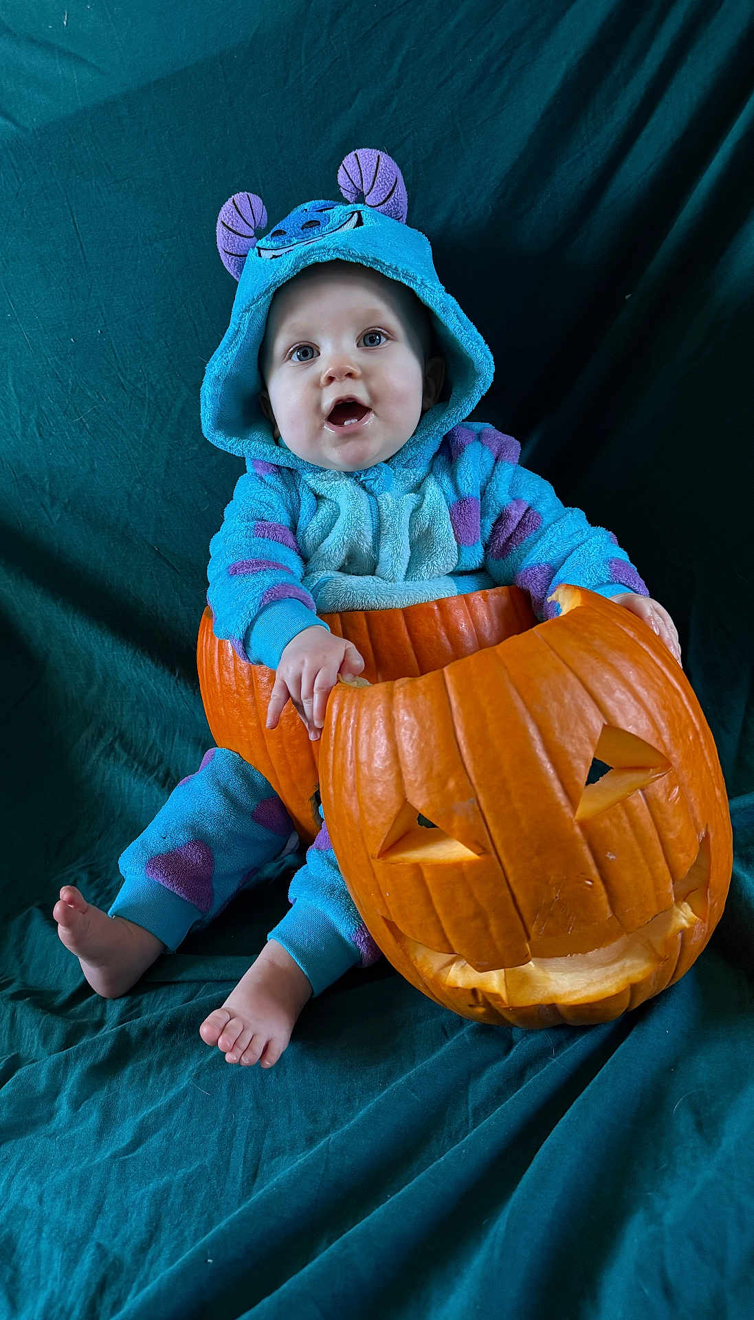 Hillarya a rejoint le concours — aidez-le/la à gagner de superbes lots ! baby, child, pumpkin, halloween, costume, onesie, blue, purple, carved_pumpkin, face, smiling, cute, sitting, barefoot, indoors, portrait, holiday, festive, dark_background, fabric
