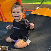 Naila is registered to the contest to win money with this photo: toddler, child, smiling, playground, swing, black_shirt, outdoor, grass, orange_equipment, happy, curly_hair, sunlight, socks, kneeling, baby, play, fun, daylight, person, casual_clothing