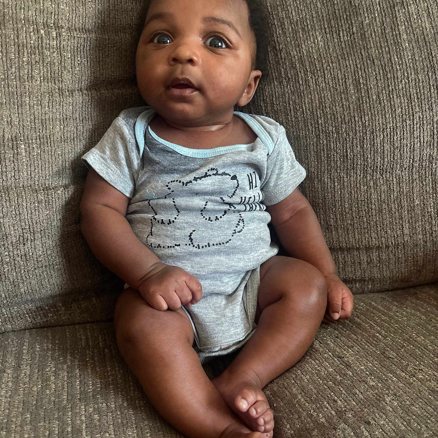 Symere joined the competition — help win amazing prizes! baby, brown, child, clothing, couch, curious, cute, expression, face, feet, furniture, hands, indoors, infant, onesie, person, portrait, sitting, skin, texture