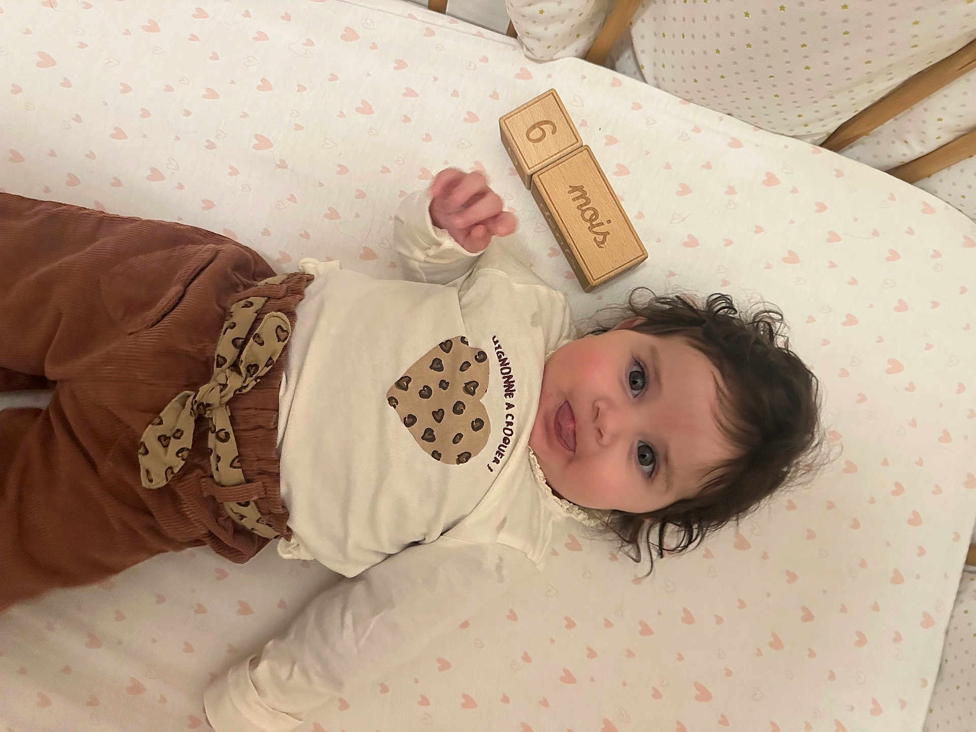 Giulia participe au concours pour gagner de l'argent avec cette photo : baby, infant, child, curly_hair, wooden_blocks, 6_months, heart_pattern, mattress, crib, brown_pants, cream_shirt, heart_design, lying_down, indoors, cute, smiling, face, skin, hand, eyes