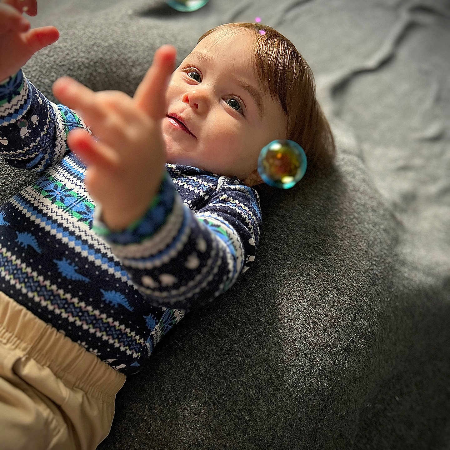 Xavier joined the competition — help win amazing prizes! toddler, child, baby, face, hands, sweater, patterned_clothing, beige_pants, bubbles, blanket, grey_background, indoor, portrait, happy, curly_hair, playful, close_up, soft_light, focus, smiling