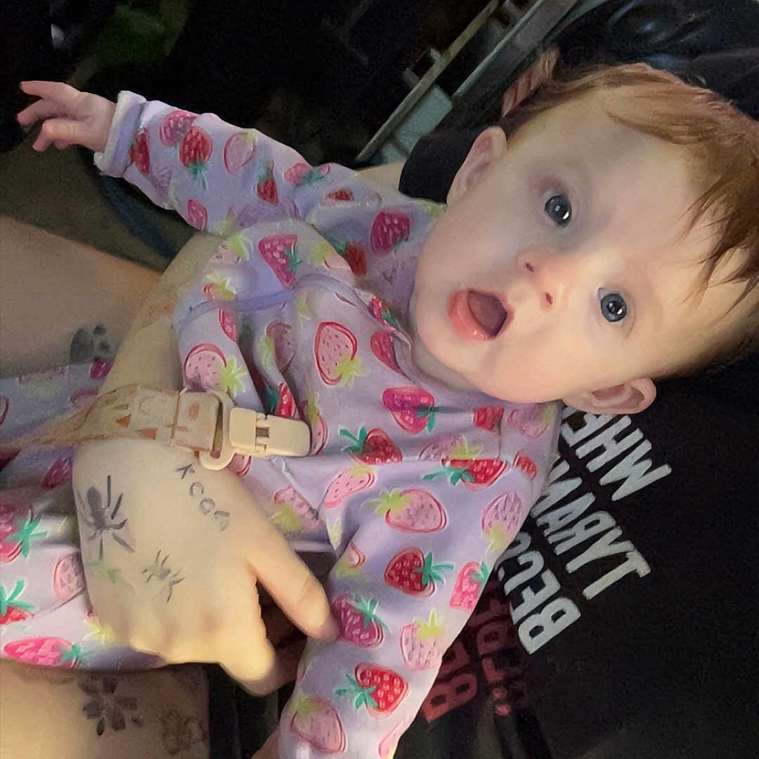 Oakley is registered to the contest to win money with this photo: baby, infant, strawberry_pattern, clothing, person, tattoo, hand, adult, holding, face, expression, wide_eyes, open_mouth, indoor, dim_lighting, curious, skin, sleeve, portrait, candid