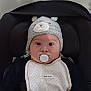 baby, infant, pacifier, hat, knitted_hat, bib, car_seat, indoor, cute, child, face, person, clothing, black_clothing, white_bib, expression, looking_at_camera, adorable, seated, headwear