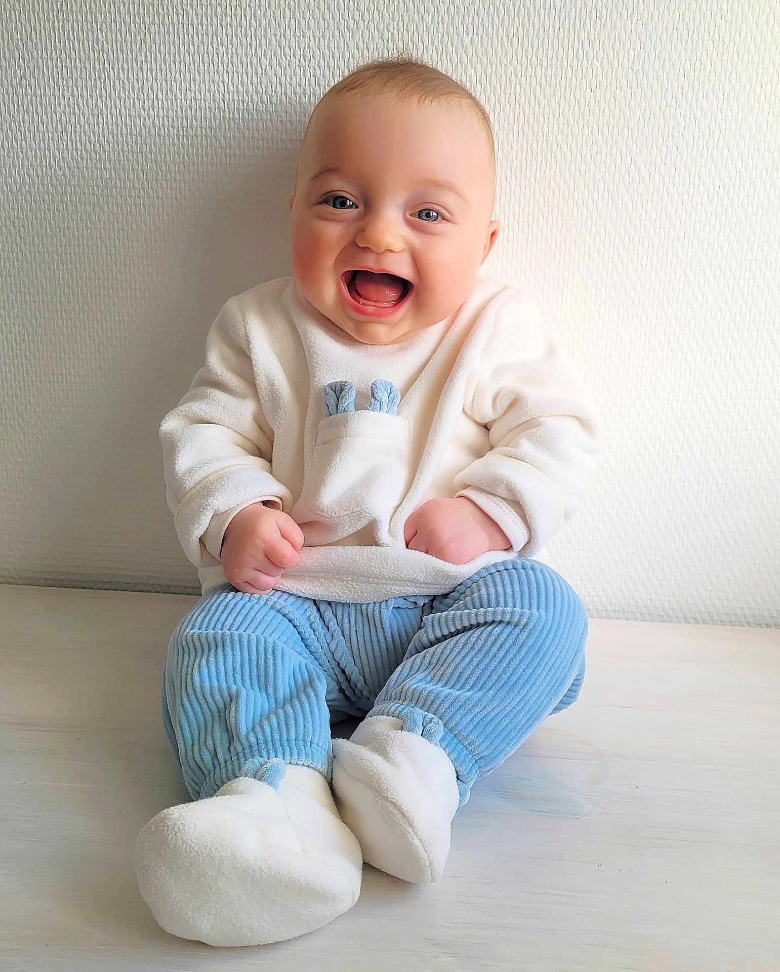 Eliott participe au concours pour gagner de l'argent avec cette photo : baby, smiling, child, sitting, blue_pants, white_sweater, indoor, floor, happy, cute, portrait, infant, warm_clothing, soft_texture, footwear, light, expression, face, hands, casual
