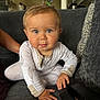 baby, blonde_hair, blue_eyes, child, couch, crawling, curious_expression, face, gray_couch, hand, indoor, infant, living_room, onesie, patterned_pillow, person, pillow, small_hands, soft_lighting, white_clothing