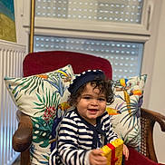 Yanis Bouasla a rejoint le concours — aidez-le/la à gagner de superbes lots ! toddler, child, striped_clothing, hat, gift_box, chair, cushion, tropical_print, indoor, smiling, curly_hair, red_chair, wooden_armrest, window, radiator, decor, happy, portrait, seated, playful