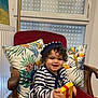 toddler, child, striped_clothing, hat, gift_box, chair, cushion, tropical_print, indoor, smiling, curly_hair, red_chair, wooden_armrest, window, radiator, decor, happy, portrait, seated, playful