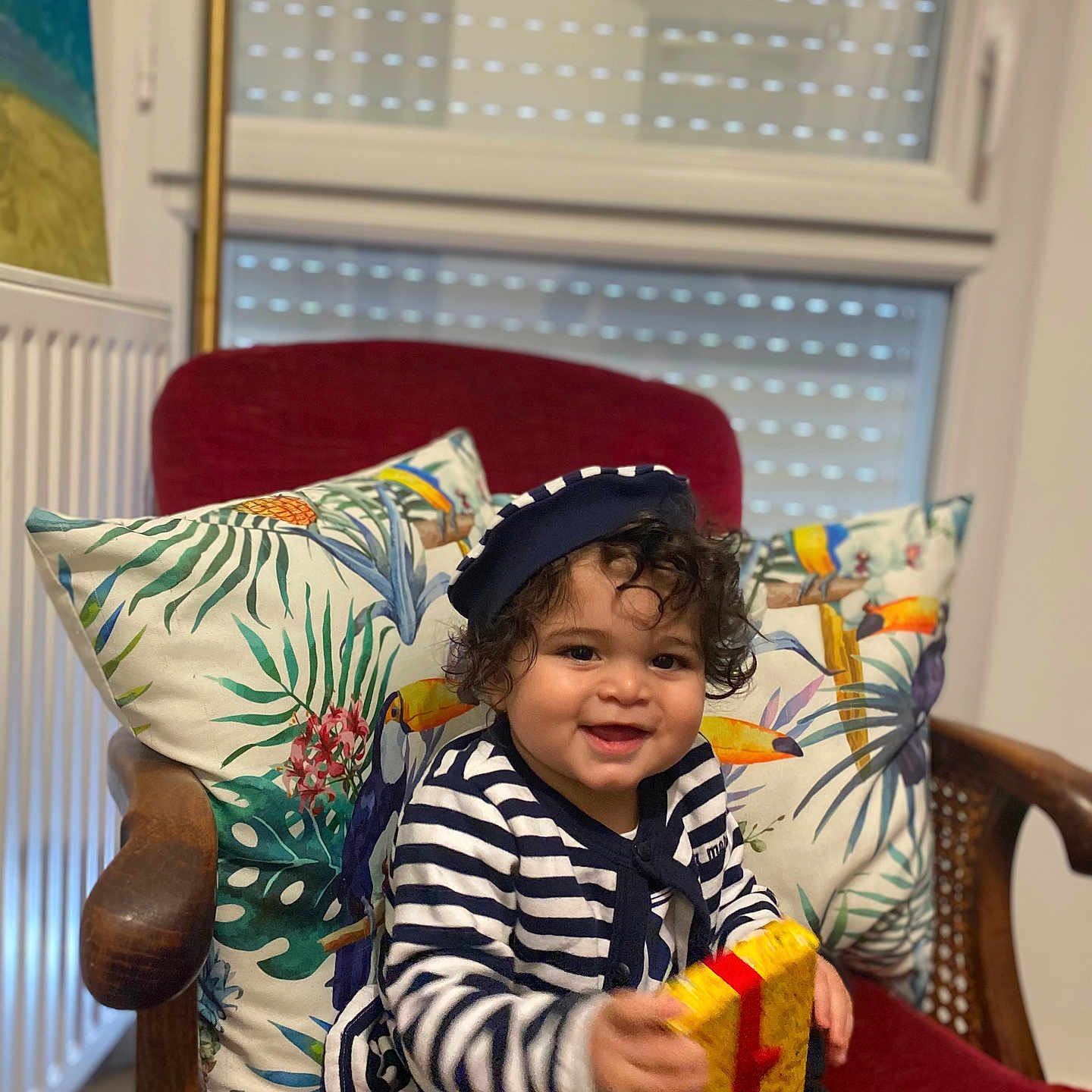 Yanis Bouasla a rejoint le concours — aidez-le/la à gagner de superbes lots ! chair, child, curly_hair, cushion, decor, gift_box, happy, hat, indoor, playful, portrait, radiator, red_chair, seated, smiling, striped_clothing, toddler, tropical_print, window, wooden_armrest