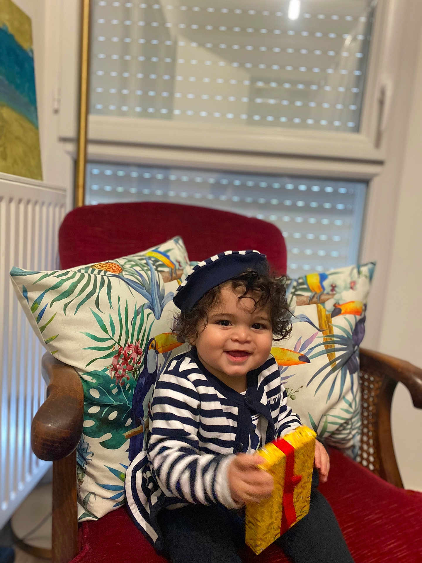 Yanis Bouasla a rejoint le concours — aidez-le/la à gagner de superbes lots ! toddler, child, striped_clothing, hat, gift_box, chair, cushion, tropical_print, indoor, smiling, curly_hair, red_chair, wooden_armrest, window, radiator, decor, happy, portrait, seated, playful