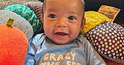 Elijah is registered to the contest to win money with this photo: baby, blue_clothing, carpet, child, colorful, cozy, cushions, cute, face, happy, home, indoor, infant, person, plush_toys, portrait, seated, smiling, soft_toys, young