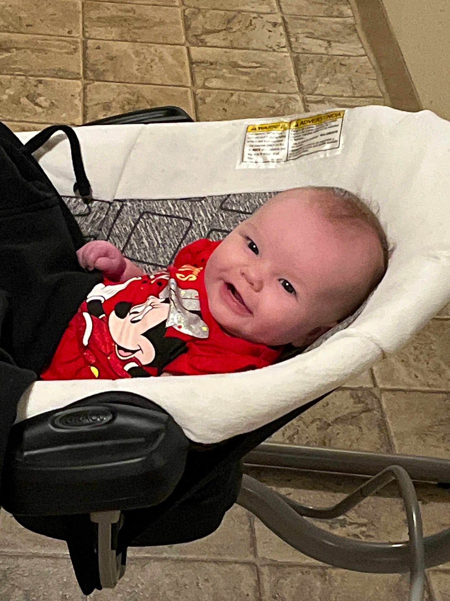 Violet is registered to the contest to win money with this photo: baby, infant, smile, red_shirt, mickey_mouse, baby_rocker, indoor, floor_tiles, happy, child, cute, person, young, clothing, seat, head, face, relaxing, home, comfort
