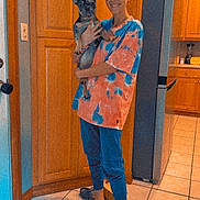 Maggie May is registered to the contest to win money with this photo: person, dog, french_bulldog, glasses, tie_dye_shirt, kitchen, wooden_cabinets, tile_floor, refrigerator, footwear, sandals, indoors, pet, standing, smiling, holding, curious, ears, home, casual