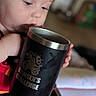 blurry_background, book, child, closeup, clothing, cup, curious, expression, face, focus, furniture, hand, holding, indoor, mug, portrait, red_shirt, sitting, skin, toddler