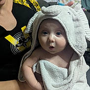 Remington is registered to the contest to win money with this photo: baby, towel, elephant_hood, surprised_expression, adult, smiling, indoor, couch, blanket, clothing, person, home, comfort, cute, child, face, portrait, sitting, cozy, holding