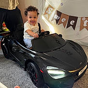Karter joined the competition — help win amazing prizes! child, toddler, toy_car, indoor, smiling, curly_hair, car, black, steering_wheel, banner, celebration, decorations, person, floor, carpet, clothing, foot, happy, party, home