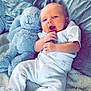 baby, plush_toy, blanket, soft_fabric, infant, cute, blue, stuffed_animal, cozy, child, person, home, comfort, relaxed, sleepwear, footed_pajamas, face, head, lying_down, indoor