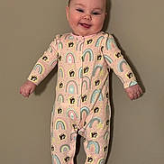 Millie is registered to the contest to win money with this photo: baby, child, pajamas, rainbow_pattern, dog_pattern, smiling, standing, indoors, face, cute, happy, onesie, footed_pajamas, infant, person, clothing, wall, skin, head, expression
