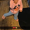toddler, child, indoor, wooden_floor, pink_shirt, blue_pants, slippers, smiling, cute, standing, hand_near_mouth, curly_hair, playful, snapchat, emoji_overlay, floor_mat, person, portrait, young_child, home