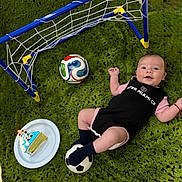 Nathan joined the competition — help win amazing prizes! baby, infant, soccer_ball, mini_soccer_goal, cake, green_rug, celebration, sports_outfit, footwear, happy, lying_down, indoor, toy, birthday, child, smiling, black_clothing, pink_clothing, plate, candle