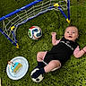 baby, infant, soccer_ball, mini_soccer_goal, cake, green_rug, celebration, sports_outfit, footwear, happy, lying_down, indoor, toy, birthday, child, smiling, black_clothing, pink_clothing, plate, candle