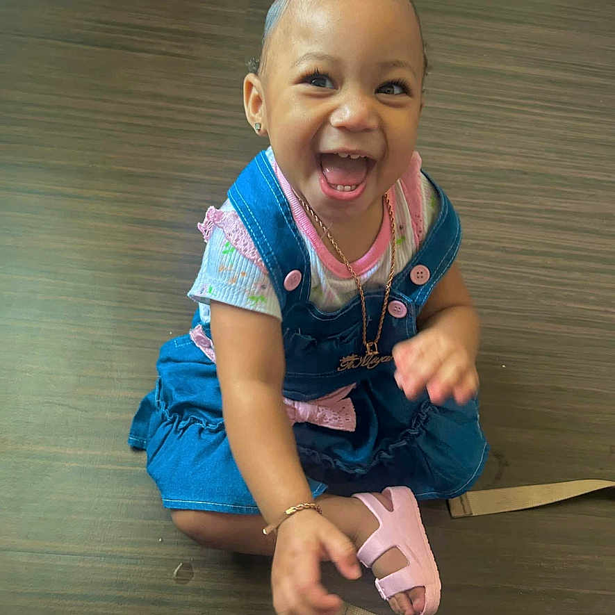Timiyah joined the competition — help win amazing prizes! baby, bracelet, child, cute, denim_dress, expression, floor, happy, indoor, jewelry, necklace, person, pink_bow, pink_sandals, playful, sitting, smiling, toddler, wooden_floor, young_child