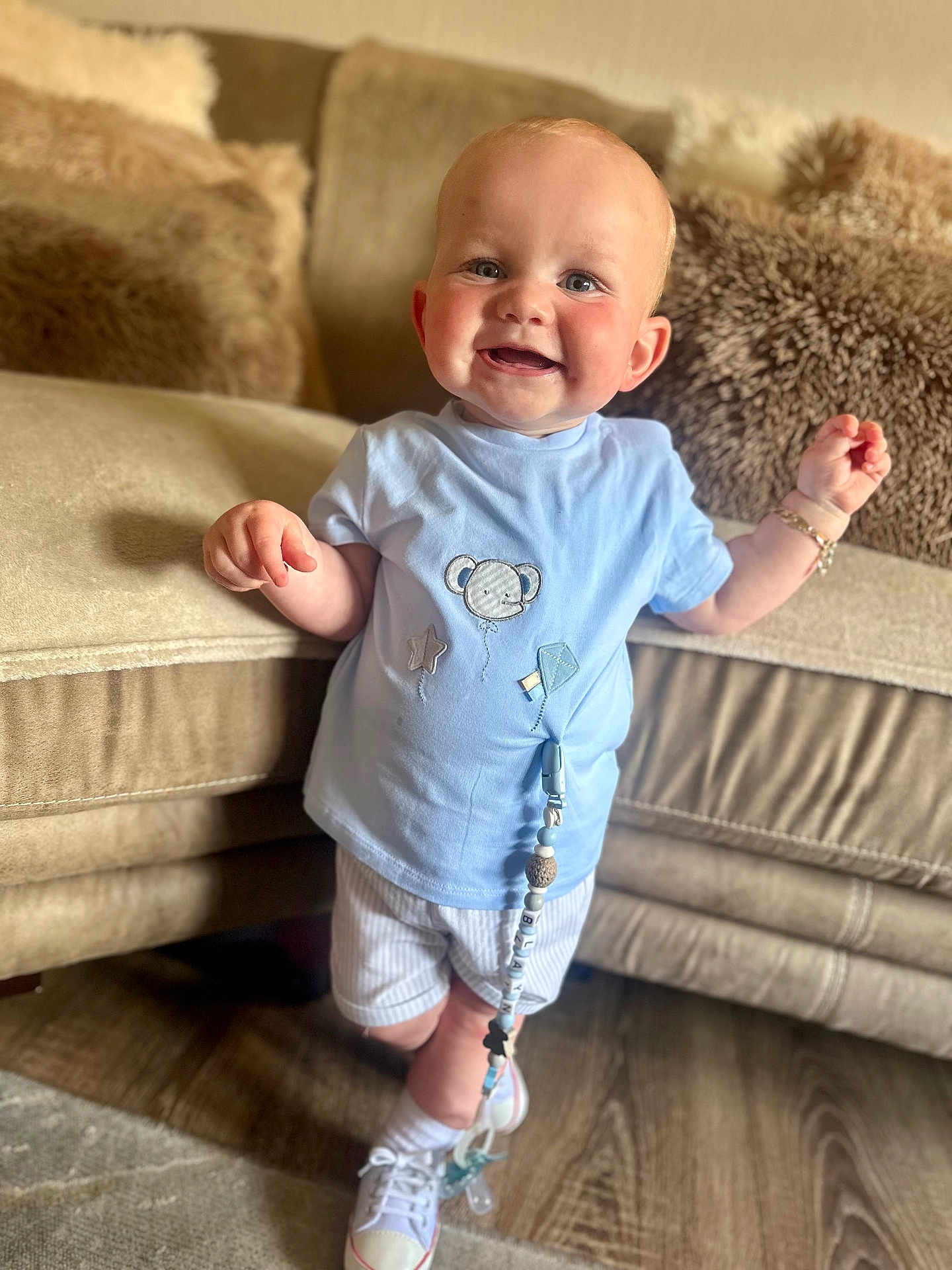 Blayn joined the competition — help win amazing prizes! baby, child, smiling, blue_shirt, shorts, white_sneakers, pacifier_clip, living_room, couch, pillows, wooden_floor, happy, standing, cute, indoor, person, face, plush, home, cozy