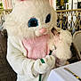 person_in_costume, bunny_costume, dog, poodle, leash, outdoor, cafe, table, chair, wicker_furniture, white_fur, pink, cute, animal, pet, affection, interaction, costume, fluffy, daylight