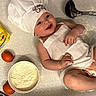 Romy a rejoint le concours — aidez-le/la à gagner de superbes lots ! baby, infant, chef_hat, apron, smiling, eggs, flour, baking, spatula, ladle, kitchen, cooking, cooking_utensils, patterned_surface, diaper, cute, happy, food_preparation, child, laying_down
