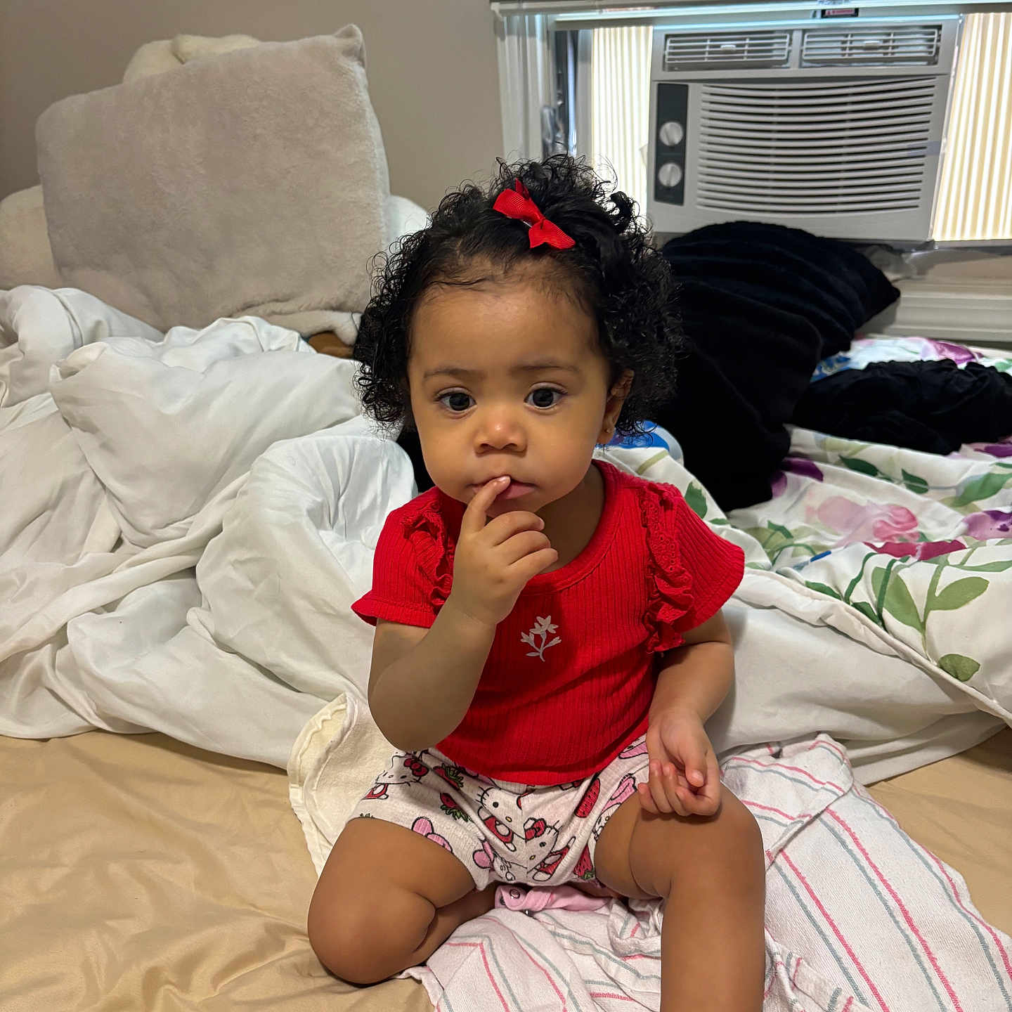 Amary is registered to the contest to win money with this photo: toddler, child, baby, red_shirt, bow, curly_hair, bed, blanket, pillow, window, air_conditioner, indoor, person, sitting, thinking, cute, home, cozy, young_child, portrait