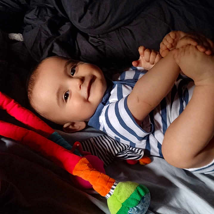 ében participe au concours pour gagner de l'argent avec cette photo : baby, bed, bedding, child, colorful_toy, cozy, cute, dark_bedding, foot_holding, happy, indoors, infant, lying_down, person, playful, plush_toy, smiling, socks, striped_onesie, young_child