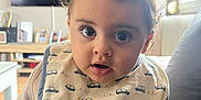 Emilio a rejoint le concours — aidez-le/la à gagner de superbes lots ! baby, child, bib, face, curly_hair, indoor, living_room, window, soft_light, clothing, person, couch, furniture, expression, portrait, cute, curious, infant, sitting, home