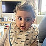 baby, child, bib, face, curly_hair, indoor, living_room, window, soft_light, clothing, person, couch, furniture, expression, portrait, cute, curious, infant, sitting, home