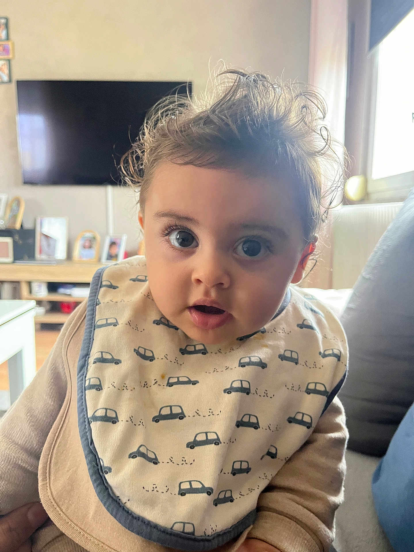 Emilio a rejoint le concours — aidez-le/la à gagner de superbes lots ! baby, child, bib, face, curly_hair, indoor, living_room, window, soft_light, clothing, person, couch, furniture, expression, portrait, cute, curious, infant, sitting, home