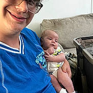 Steven joined the competition — help win amazing prizes! baby, adult, blue_shirt, glasses, couch, infant, person, indoor, smiling, holding, onesie, baby_crib, fabric, casual, home, comfort, relaxed, seated, portrait, furniture