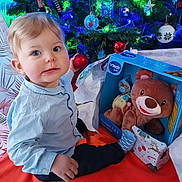 Yaël a rejoint le concours — aidez-le/la à gagner de superbes lots ! blue_shirt, celebration, child, christmas_tree, decorations, festive, floor, gift_box, holiday, indoor, ornament, person, pine_tree, present, smile, sock, teddy_bear, toddler, toy, wrapping_paper