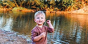 Oliver is registered to the contest to win money with this photo: child, boy, blond_hair, blue_eyes, river, water, trees, nature, outdoor, sweater, pebble, shore, smile, young, daylight, forest, reflection, casual_clothing, happy, playful