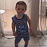 bare_legs, blue_and_white, child, clogs, curtains, home_interior, indoor, neutral_expression, one_person, pacifier, portrait, short_hair, sliding_door, socks, standing, striped_outfit, tile_floor, toddler, window, young_child