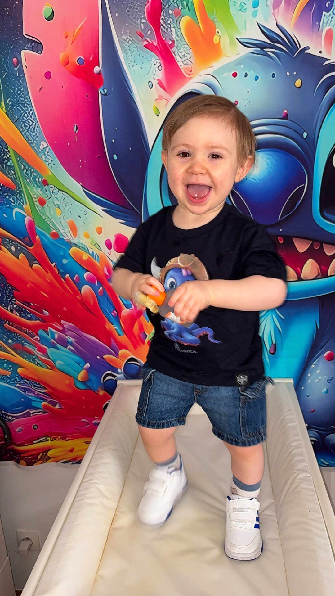 Thélyo a rejoint le concours — aidez-le/la à gagner de superbes lots ! child, toddler, smiling, standing, toy, sneakers, denim_shorts, tshirt, colorful_background, wall_art, cartoon_character, portrait, happy, indoor, playful, cheerful, face, hands, eyes, mouth