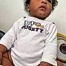 baby, child, curly_hair, white_shirt, text_on_clothing, lion_graphic, plush_toy, eye_graphic, indoor, soft_lighting, cute, innocence, curiosity, portrait, seated, close_up, hand, skin, face, clothing